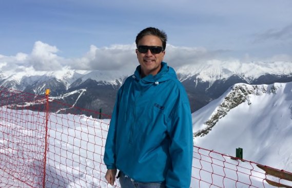 Bruce Kaplan standing in a blue ski jacket at the top of the resort, snow-covered peaks behind him
